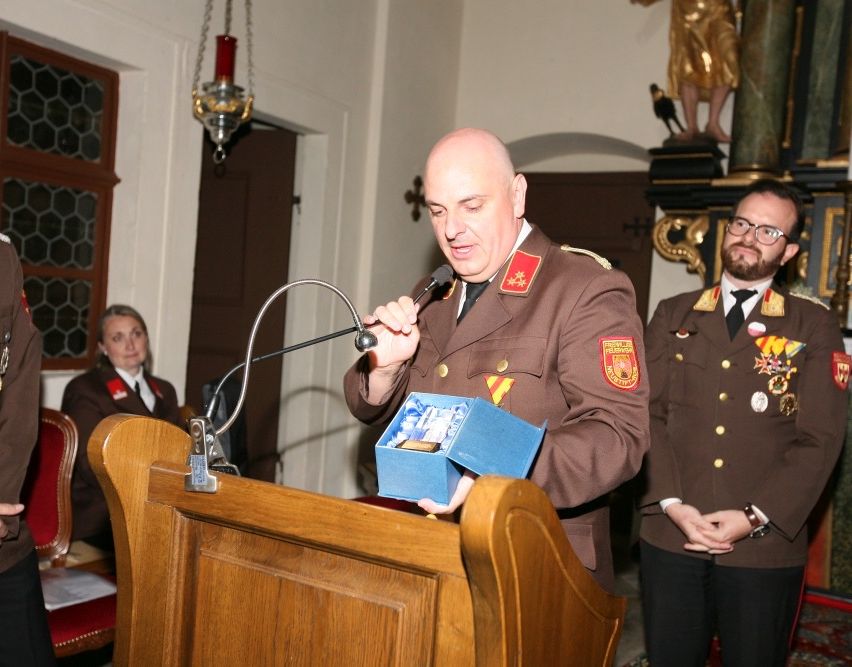 Ein Mann in Uniform hält eine blaue Schachtel mit einem Band und spricht in ein Mikrofon am Rednerpult.