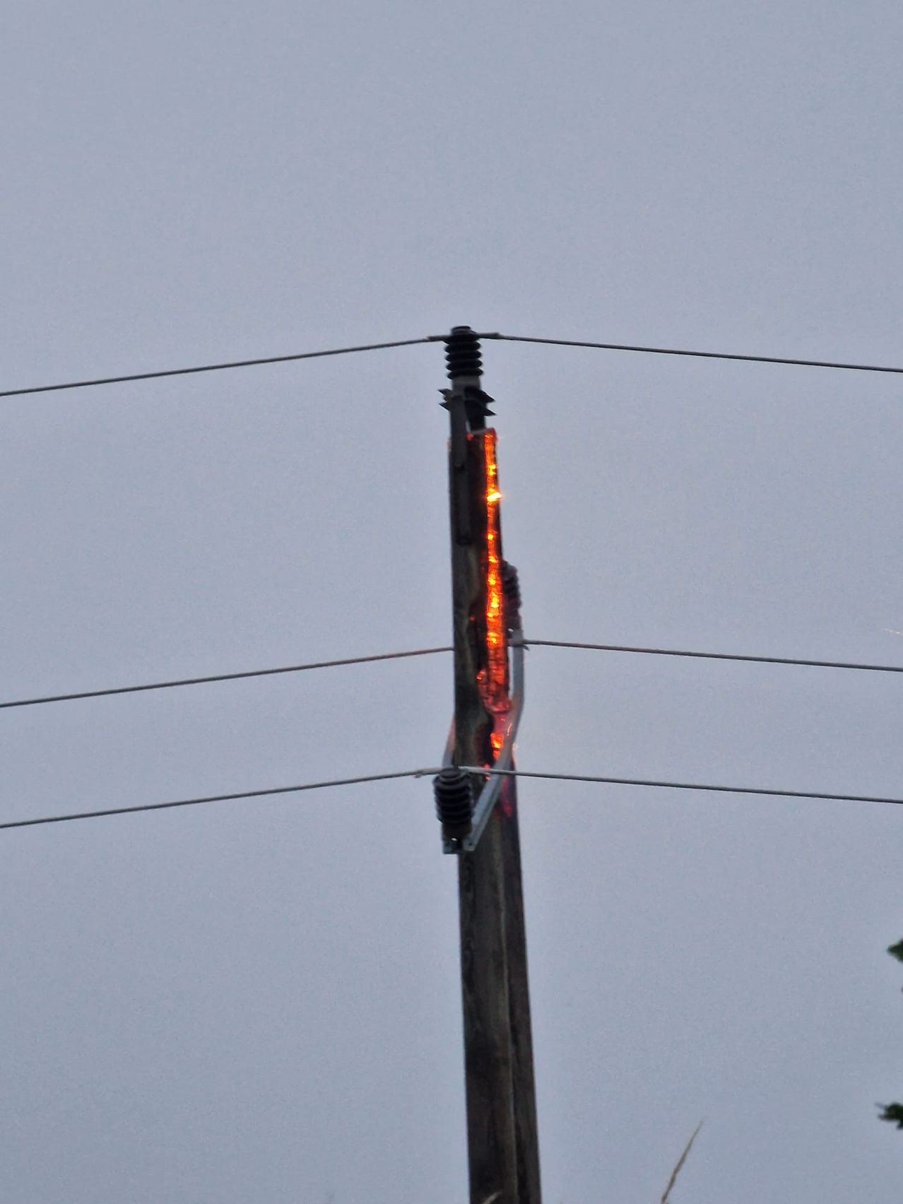 Ein Strommast zeigt Anzeichen von Schäden, wobei Flammen an seiner Seite unter einem grauen Himmel sichtbar sind.