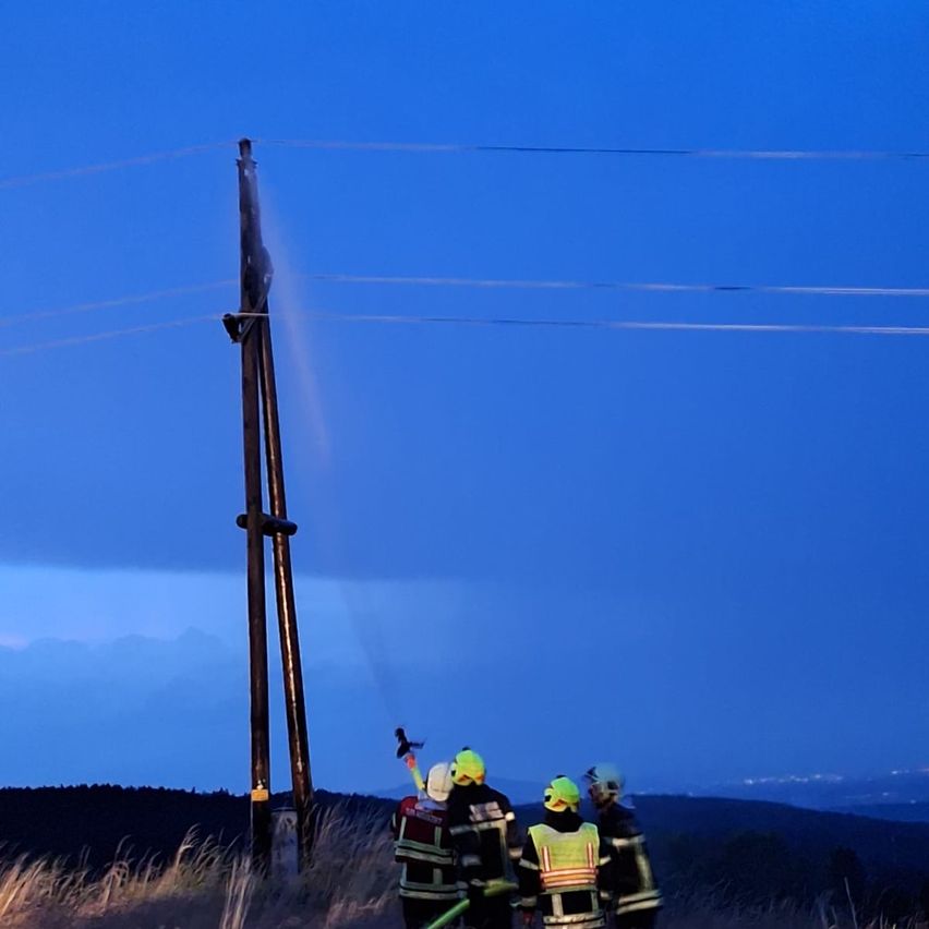 Drei Feuerwehrleute stehen neben einem Strommast mit Stromleitungen in einem ländlichen Gebiet bei Einbruch der Dunkelheit. Sie tragen Sicherheitswesten und Helme.