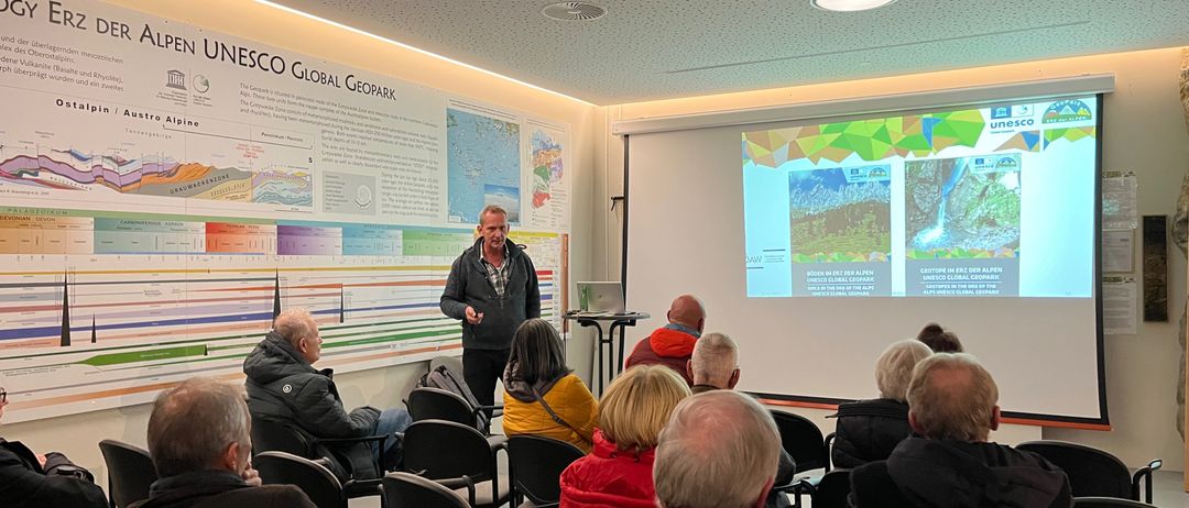 Ein Mann hält eine Präsentation vor einer Projektionsleinwand in einem Konferenzraum. Mehrere Personen sitzen auf Stühlen und lauschen. Die Projektionsleinwand zeigt eine Karte.