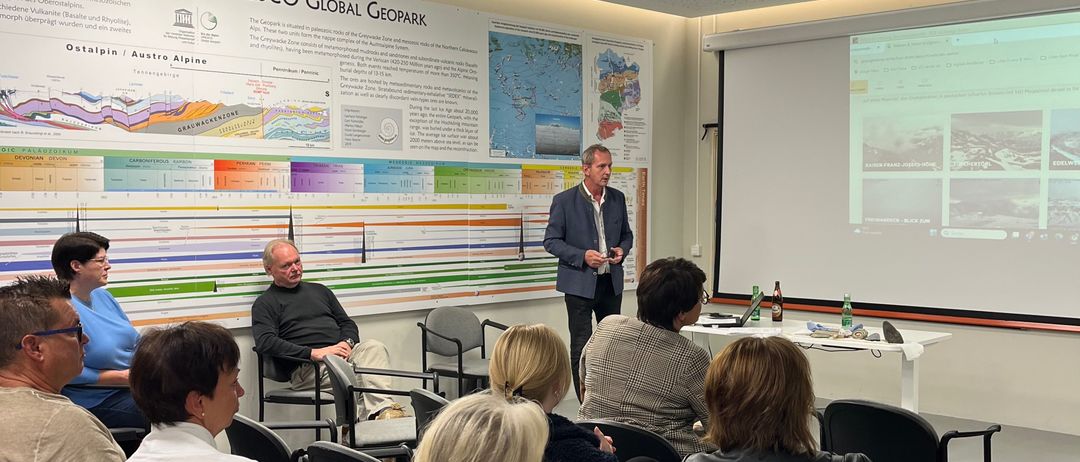 Ein Mann hält eine Präsentation in einem Konferenzraum mit einer Projektionsleinwand und Teilnehmern. Ein Banner mit dem Text 'UNESCO Global Geopark' ist an der Wand angebracht.