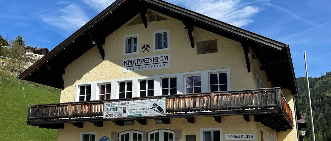 Ein gelbes Gebäude mit Balkon und mehreren Fenstern steht vor einem grünen Hügel. Das Gebäude trägt den Namen Knäppenheim. Draußen sind Bänke.