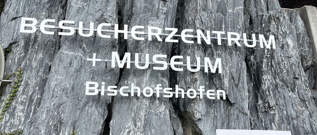 Eine große Felswand mit weißem Text, der 'BESUCHERZENTRUM + MUSEUM Bischofshofen' lautet, ist zu sehen. Der Fels ist grau und hat etwas Grünpflanzen darum herum. Rechts befindet sich ein Schild mit Logos und Text.