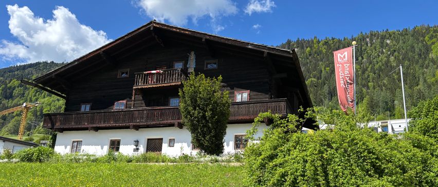 Ein traditionelles Holzhaus mit Balkon und Fahne steht auf einer Wiese mit Bergen im Hintergrund.