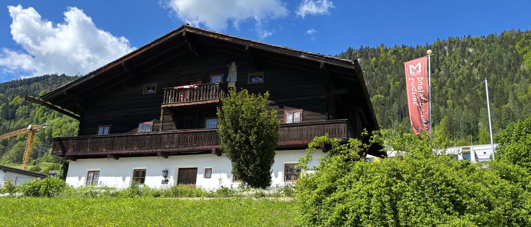 Ein traditionelles Holzhaus mit Balkon und Fahne steht auf einer Wiese mit Bergen im Hintergrund.