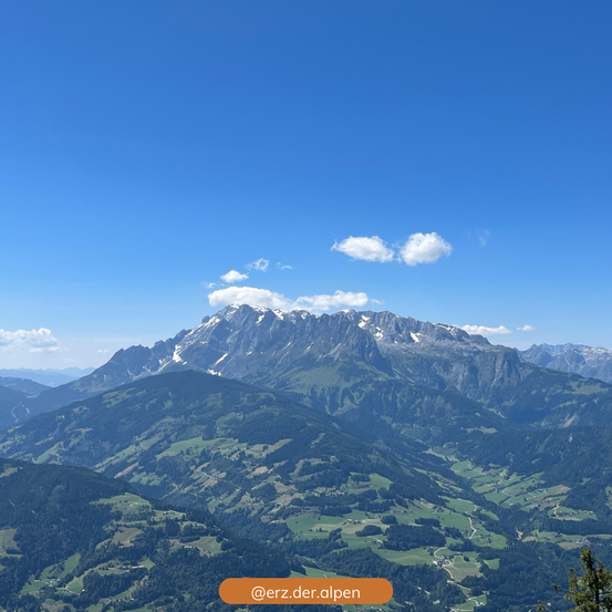 Luftaufnahme einer Bergkette unter einem klaren blauen Himmel mit ein paar Wolken. Die Berge sind mit grünen Wäldern und Wiesen bedeckt. Ein Wasserzeichen befindet sich unten.