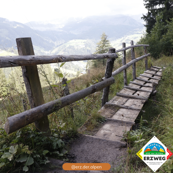 Eine Holzbrücke führt über einen Grashügel mit einem Holzzaun. Dahinter befinden sich Hügel und Bäume. Es gibt ein Logo mit Text für Erz der Alpen.