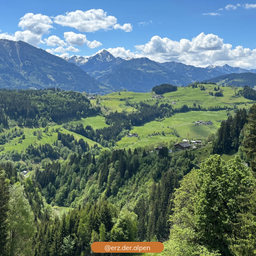 Eine landschaftliche Vogelperspektive eines grünen Tals, umgeben von hohen Bäumen und Bergen unter einem bewölkten Himmel. Ein Wasserzeichen mit @erz.der.alpen befindet sich unten.