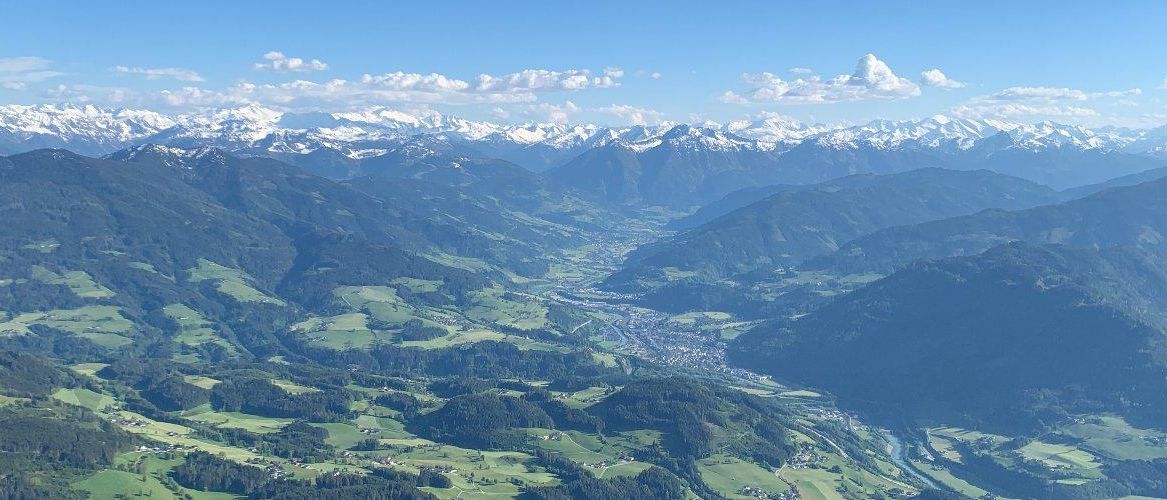 Eine hochgelegene Luftaufnahme eines Gebirgszugs mit einer Stadt darunter. Schneebedeckte Gipfel sind in der Ferne, und üppige grüne Täler umgeben sie. Der Himmel ist blau mit verstreuten Wolken.