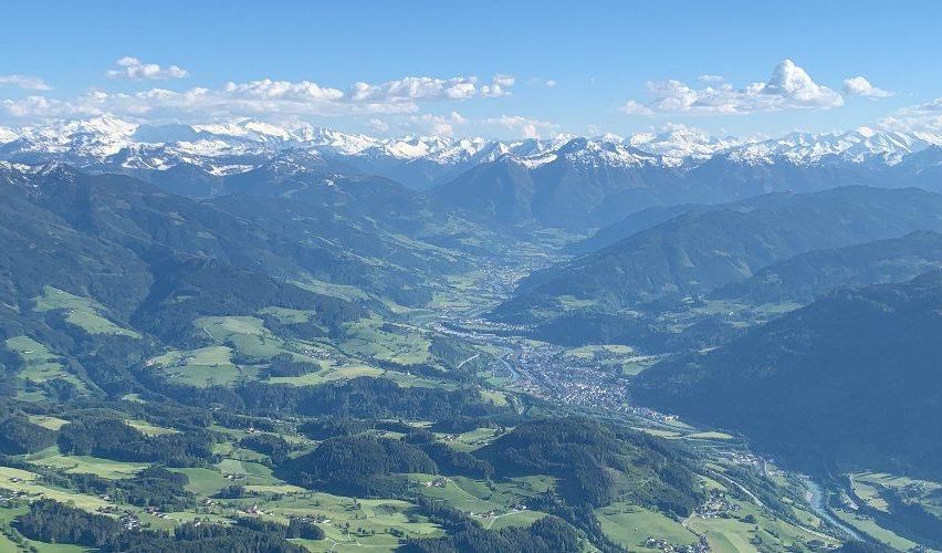Eine hochgelegene Luftaufnahme eines Gebirgszugs mit einer Stadt darunter. Schneebedeckte Gipfel sind in der Ferne, und üppige grüne Täler umgeben sie. Der Himmel ist blau mit verstreuten Wolken.