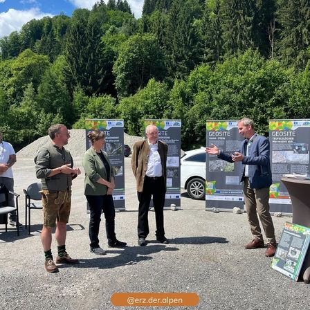Eine Gruppe von Personen steht vor Informationstafeln in einem Kiesbereich mit Bäumen und einem Auto im Hintergrund. Sie scheinen die Informationen auf den Tafeln zu diskutieren.