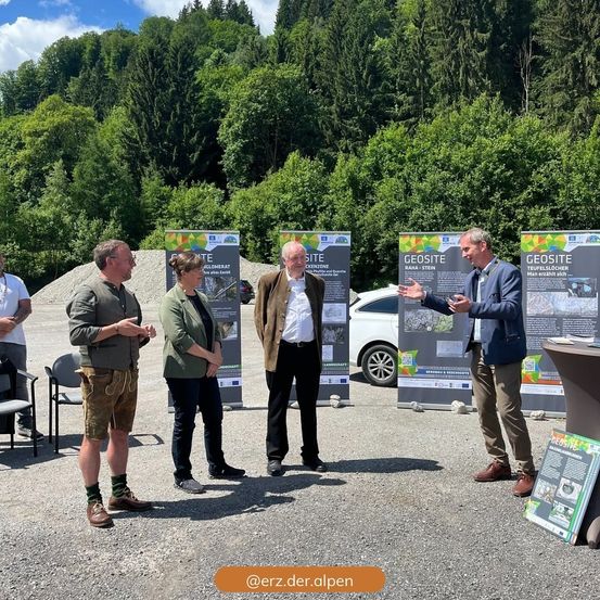 Eine Gruppe von Personen steht vor Informationstafeln in einem Kiesbereich mit Bäumen und einem Auto im Hintergrund. Sie scheinen die Informationen auf den Tafeln zu diskutieren.