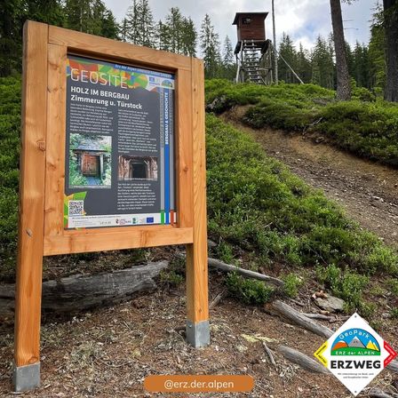 Ein Holzschild zeigt Informationen über einen GeoSite, der sich in einem Gebirgsgebiet befindet, mit einem hölzernen Aussichtsturm im Hintergrund.