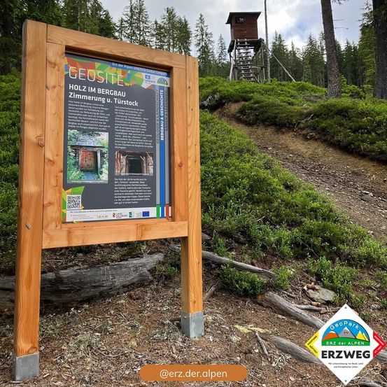 Ein Holzschild zeigt Informationen über einen GeoSite, der sich in einem Gebirgsgebiet befindet, mit einem hölzernen Aussichtsturm im Hintergrund.