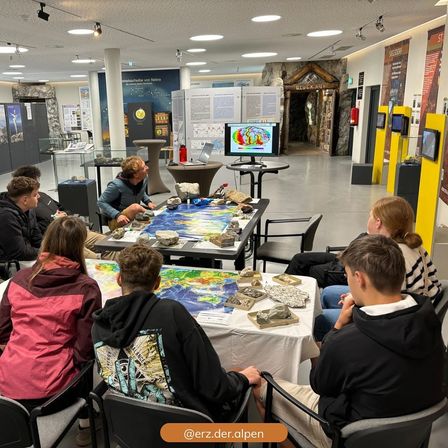 Eine Gruppe von Menschen sitzt in einem Raum um einen Tisch herum, ein Mann steht und spricht. Auf dem Tisch liegt eine Karte und ein Monitor. Der Raum hat eine gelbe Wand, einen Feuerlöscher und eine Glasvitrine.