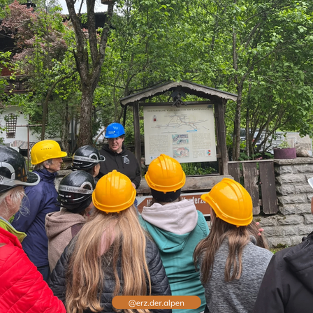 Eine Gruppe von Menschen mit Helmen steht um einen Informationspfosten in einem Park. Der Mann in der Mitte trägt einen blauen Helm und scheint etwas zu erklären.