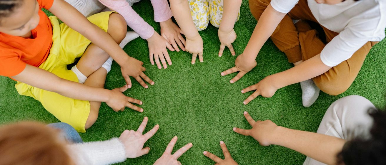 Eine Gruppe von Kindern sitzt im Kreis auf einem grünen Teppich und hält Händchen, wahrscheinlich nehmen sie an einer Gruppenaktivität oder einem Spiel teil.