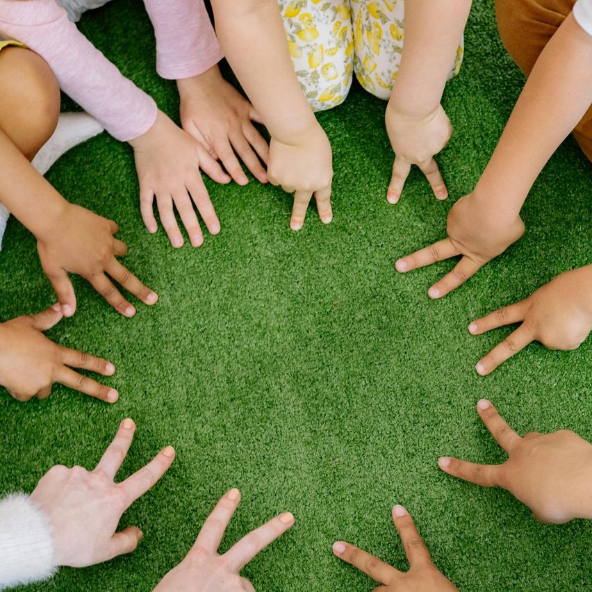 Eine Gruppe von Kindern sitzt im Kreis auf einem grünen Teppich und hält Händchen, wahrscheinlich nehmen sie an einer Gruppenaktivität oder einem Spiel teil.