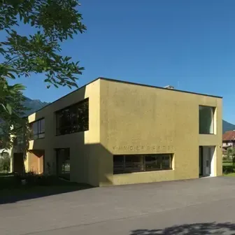 Ein gelbes Gebäude mit mehreren Fenstern ist von Bäumen und Grün umgeben. Es befindet sich in einem Parkplatz mit klarem blauem Himmel.