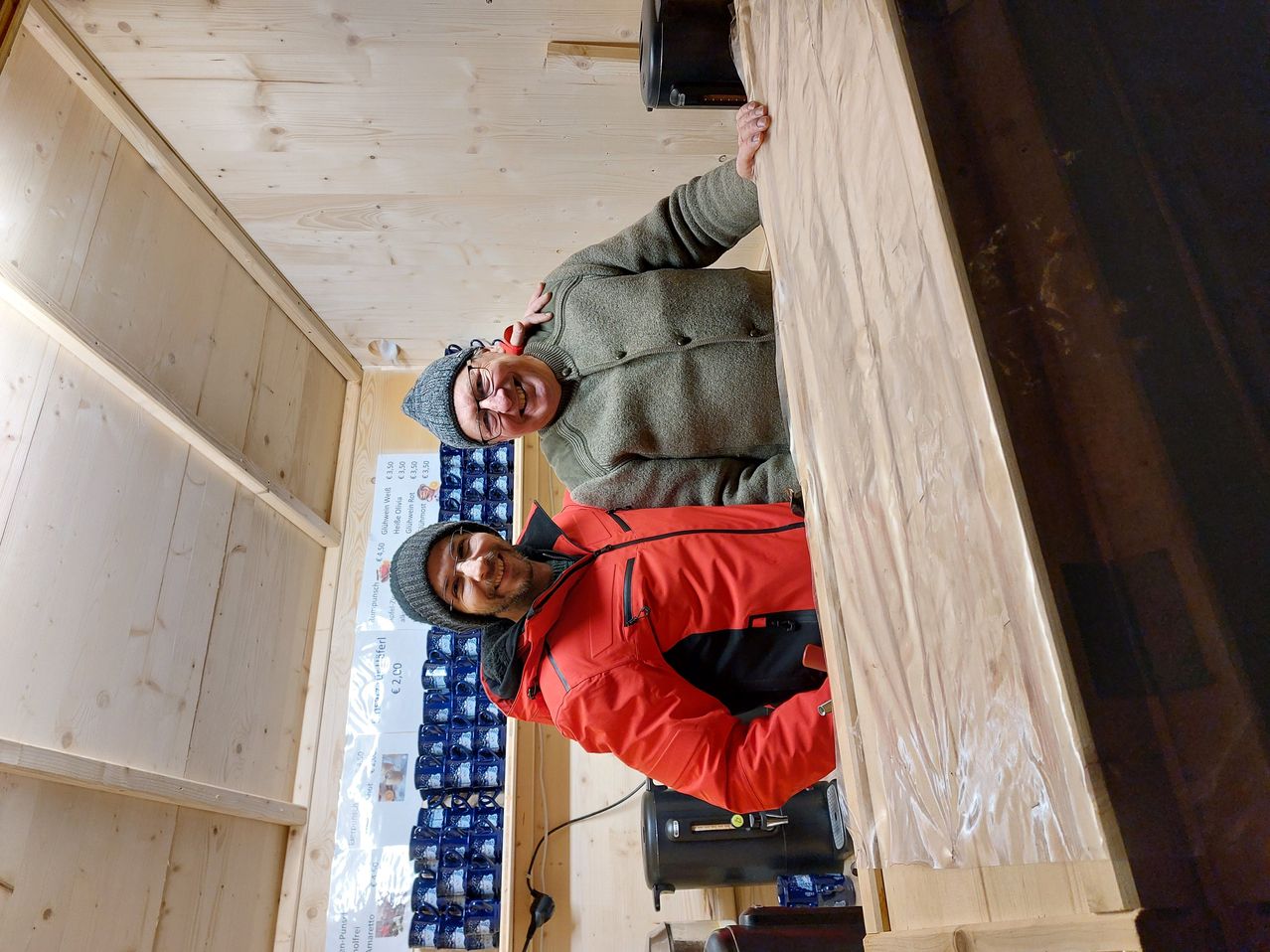 Zwei Männer stehen in einem Raum mit Holzwänden. Ein Mann lehnt an der Wand und lächelt. Der andere Mann steht neben ihm und lächelt auch. Hinter ihnen sind Tassen gestapelt und ein Poster an der Wand.