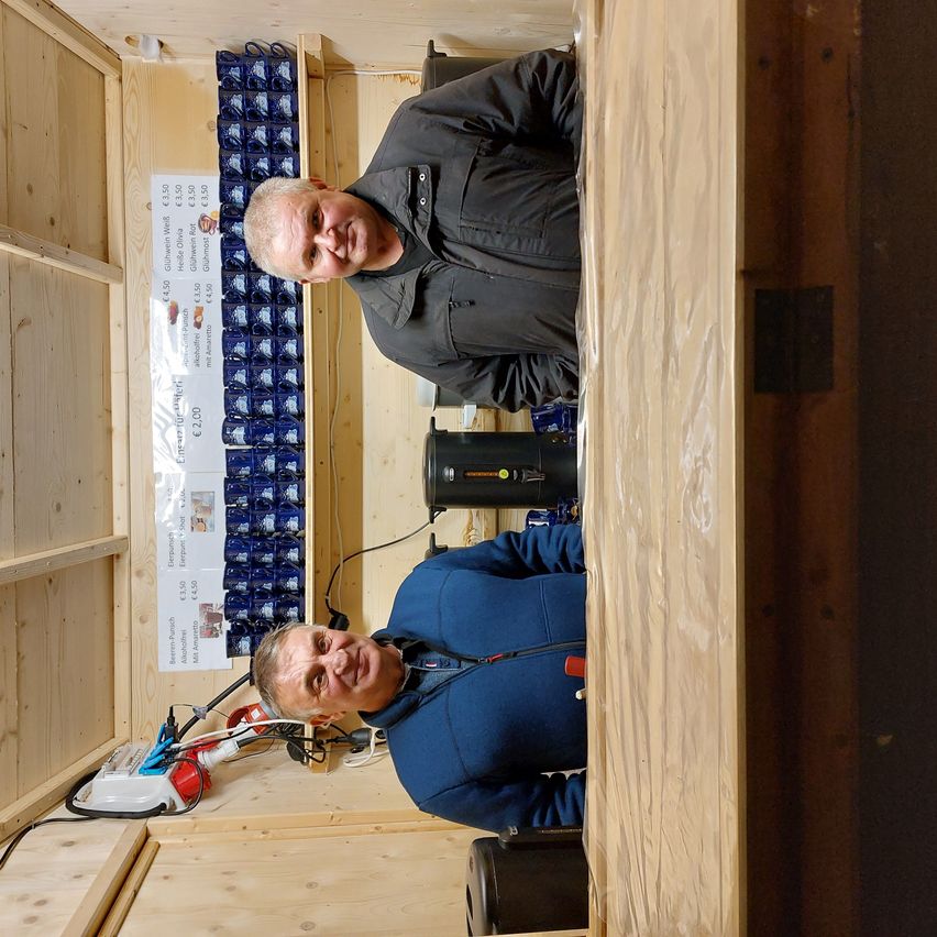 Zwei Männer in einem kleinen Raum, einer lächelt und lehnt sich an die Wand, der andere posiert lächelnd.