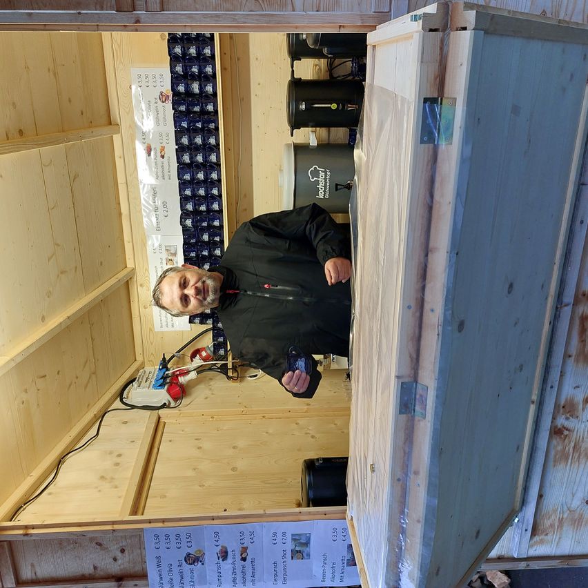 Ein Mann in einer schwarzen Jacke lehnt sich gegen eine Holzstruktur und hält ein Handy. Hinter ihm sind Regale und eine weiße Fahne zu sehen.