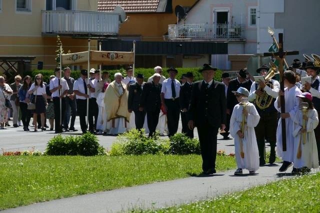 A religious procession is taking place on a street, with a cross bearer and a child in front. A priest is leading the procession, and behind him are people in formal attire.
