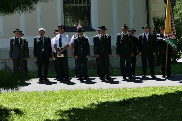A group of men in uniform stand in a row in front of a building with a flag in the right corner.