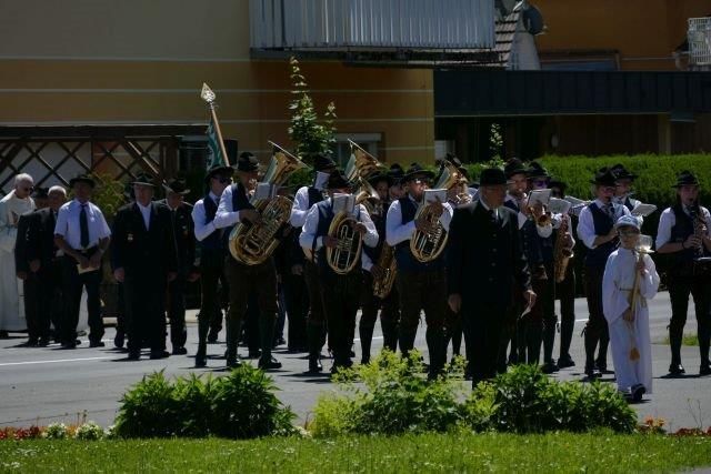 A marching band in traditional attire, holding instruments, is standing on a street. Behind them is a building with a balcony.