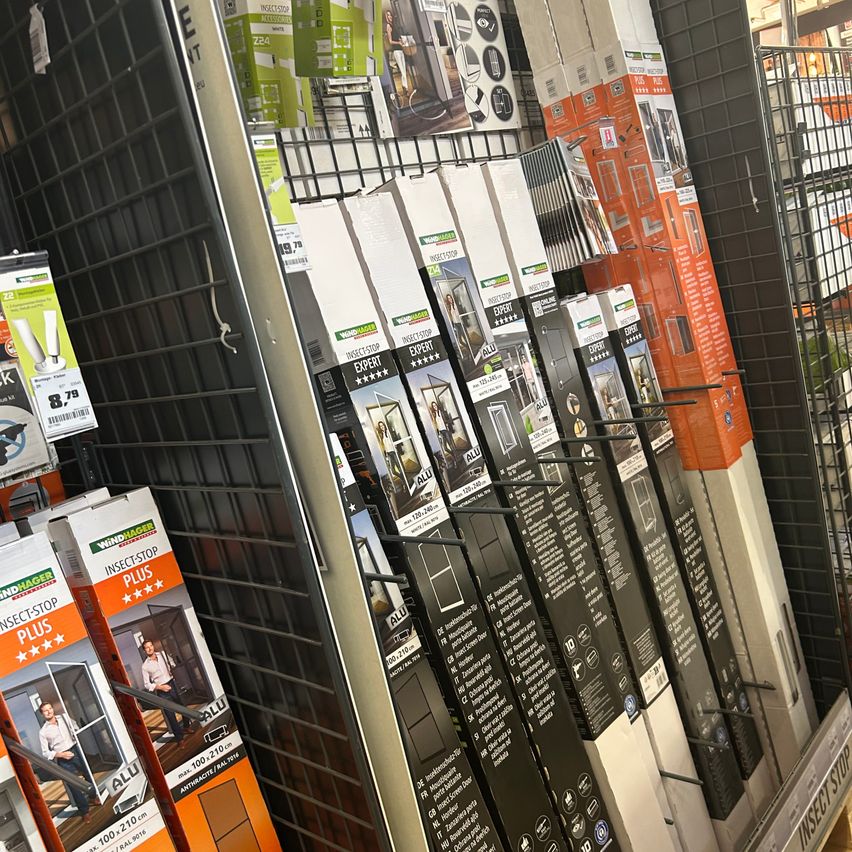 Ein Ladendisplay zeigt verschiedene Windhager-Produkte, darunter Insektenschutzgitter und Türen, alle ordentlich auf einem Metallgitter angeordnet.