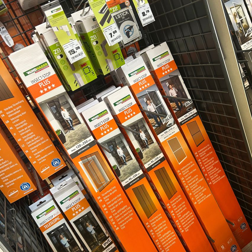 Ein Ladenregal zeigt mehrere orangefarbene und weiße Verpackungen von WindHager Insektenstop-Produkten, jeweils mit einem Mann und einer Frau vor einer Tür abgebildet.