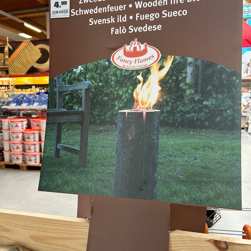 Ein Schild bewirbt Fancy Flames Holzfeuerschalen mit einem Preisschild von 4,99. Das Bild zeigt eine Feuerschale auf einem Baumstumpf mit einer Bank in der Nähe.