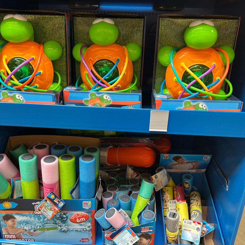 Ein Ladenregal zeigt drei farbenfrohe Schildkrötenspielzeuge, jedes mit einem zentralen Ring aus bunten Ringen, und darunter Boxen mit verschiedenen Wasserspielzeugen und Poolzubehör.