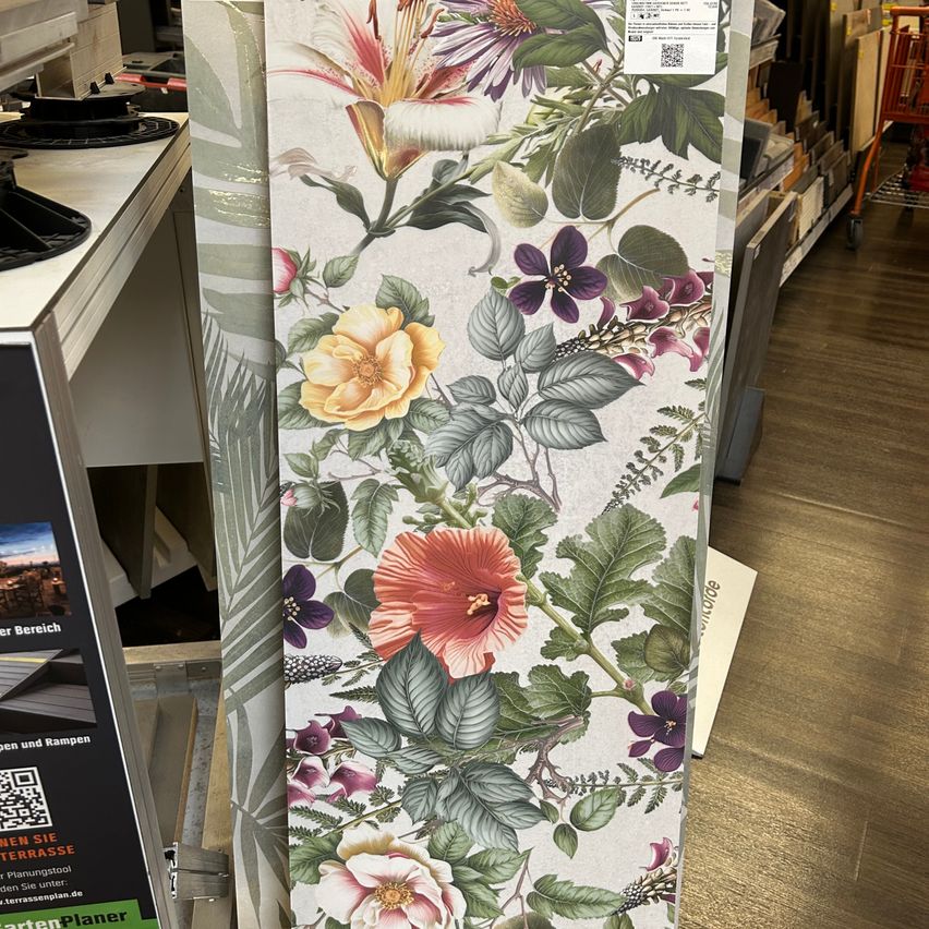 Eine florale Tapete mit einer Mischung verschiedener Blumen und Blätter wird in einem Geschäft ausgestellt, mit einem Tisch und Regalen im Hintergrund.