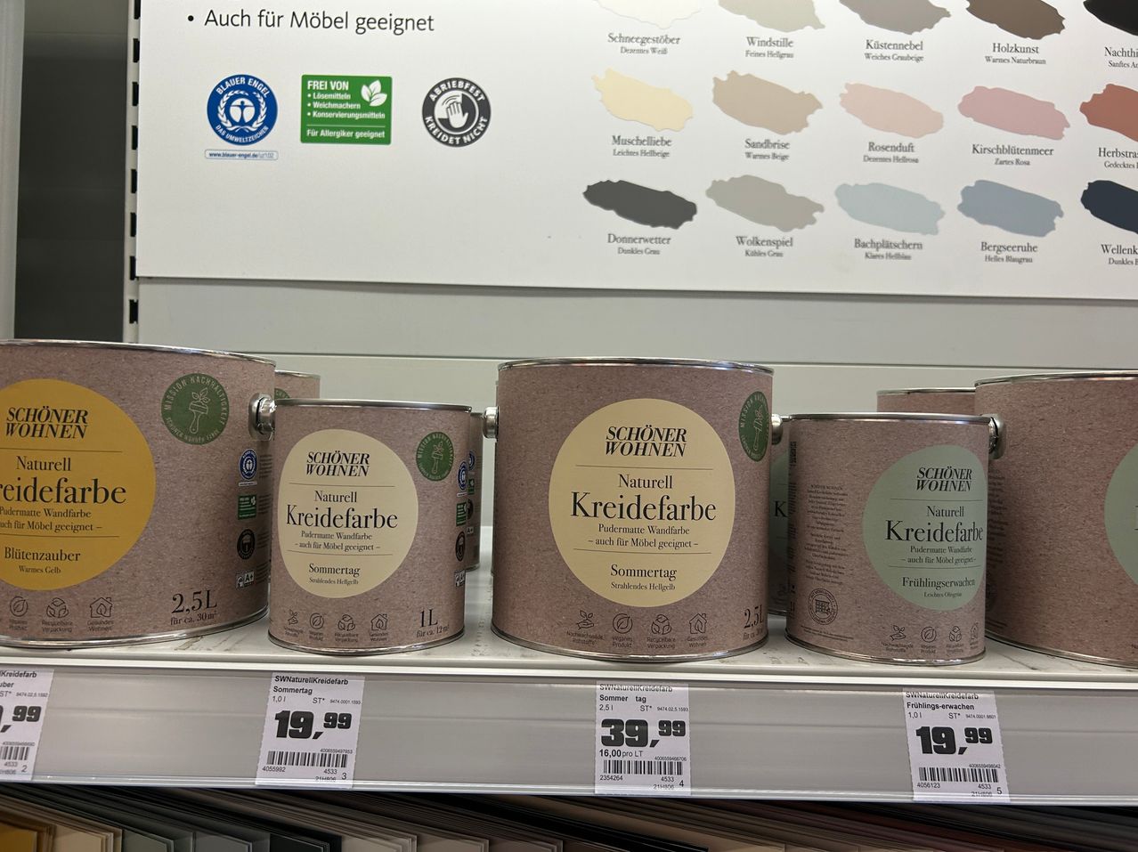 Ein Regal in einem Geschäft zeigt vier Dosen 'Schöner Wohnen' Kreidefarbe. Die Dosen sind mit verschiedenen Farben und Größen beschriftet. Ein weißer Hintergrund zeigt eine Farbpalette.