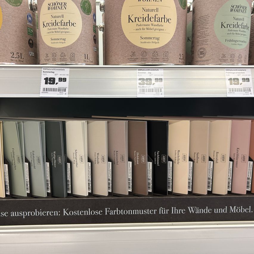 Ein Regal mit verschiedenen Farbdosen, die mit 'Kreidefarbe' und 'Schöner Wohnen' beschriftet sind. Preisschilder zeigen Preise in Euro. Unten sind Farbmuster verschiedener Farben ausgestellt.