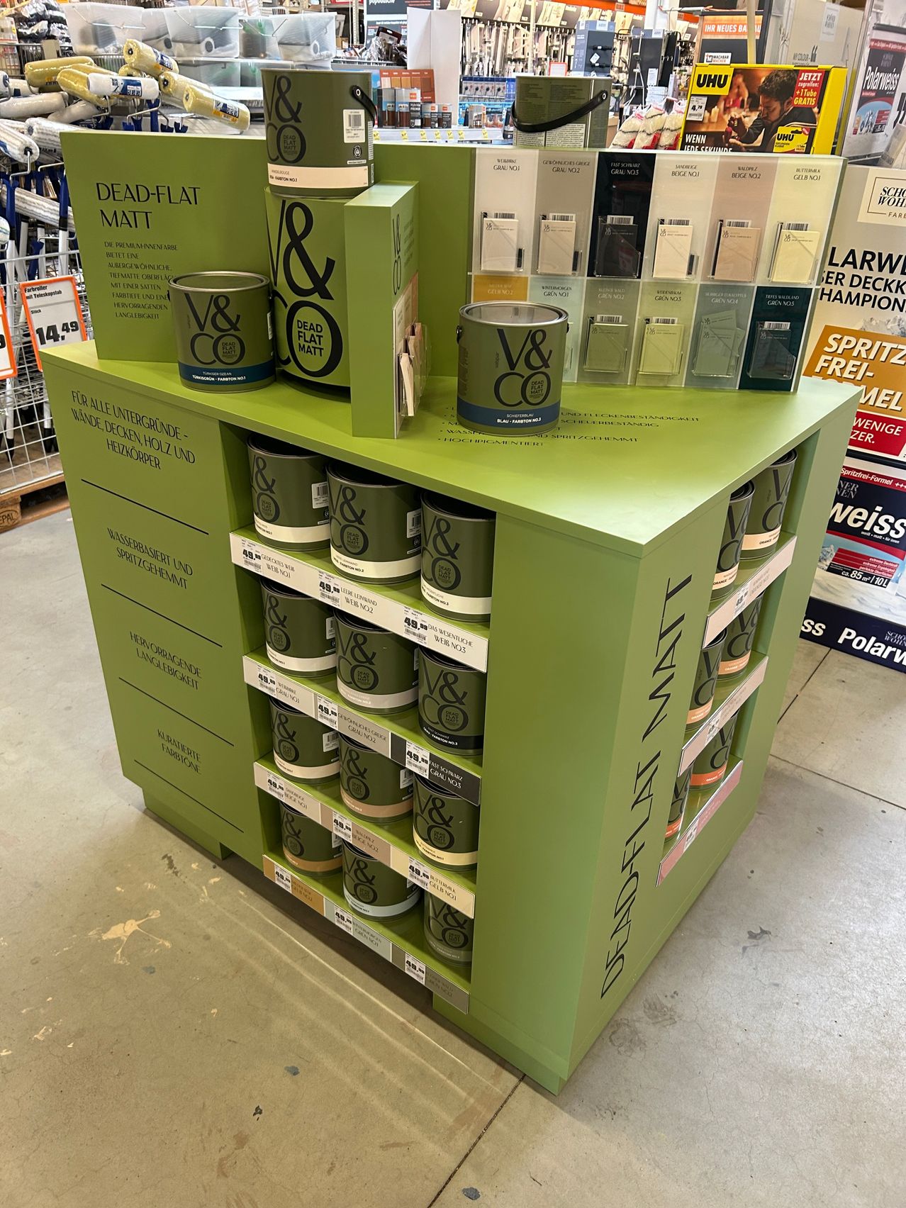 Ein Ladendisplay mit grünen Regalen, das Farbdosen mit V&CO und DEAD FLAT MATT präsentiert. Die Dosen sind in Reihen angeordnet mit Preisschildern und Werbeschildern.