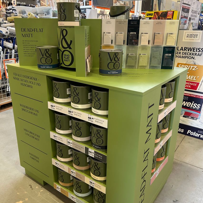 Ein Regaldisplay mit grünen Farbdosen, die mit 'Dead Flat Matt' beschriftet sind. Die Dosen sind in Reihen auf einem grünen Regal angeordnet. Zusätzliche Informationen zu den Produkten sind an der Rückwand angebracht.