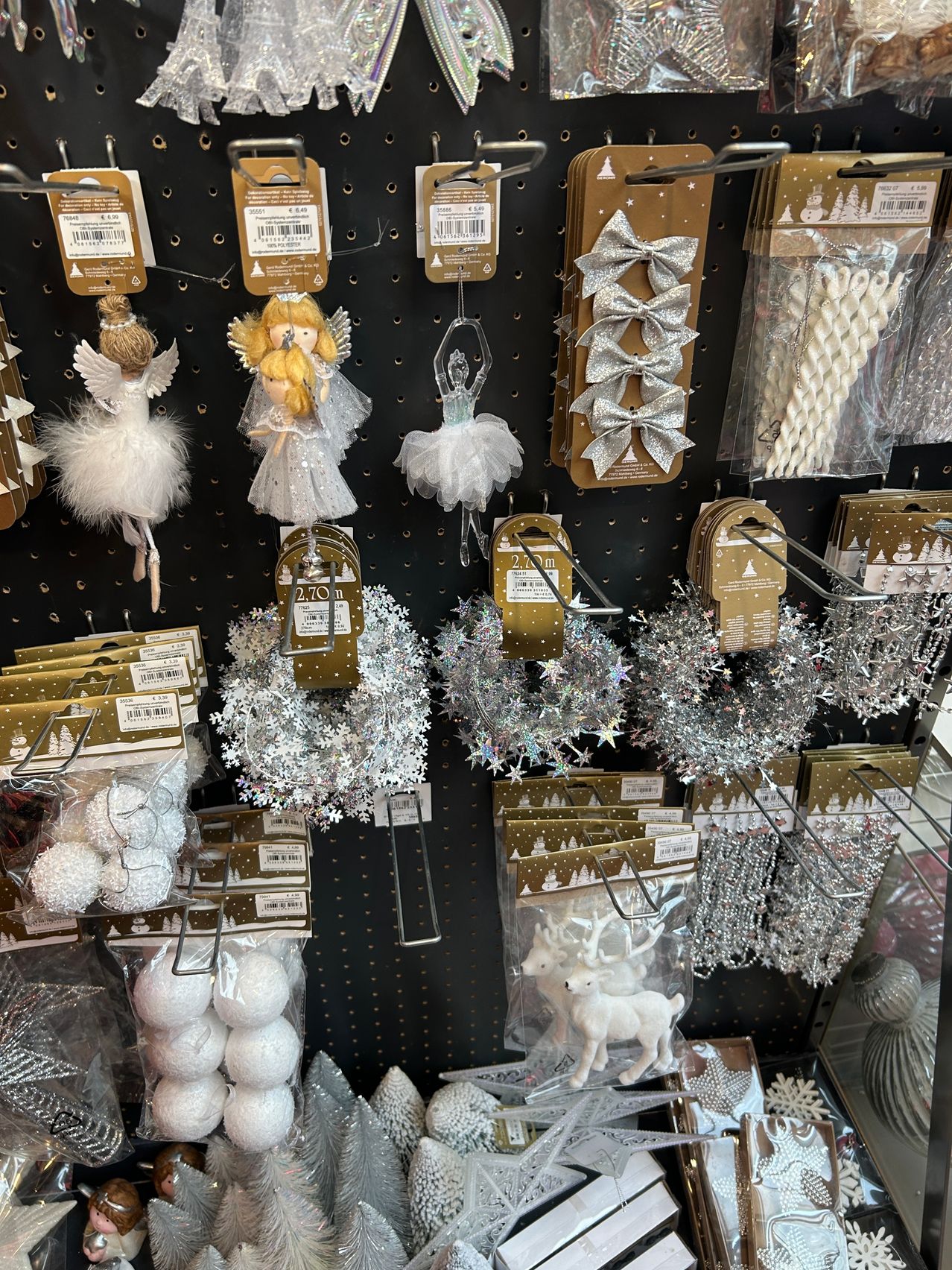 Eine Wandausstellung von Weihnachtsdekorationen, einschließlich Engel-Ornamente, Ballerinenfiguren, silberne Schleifen, Schneeflocken und Rentiere. Jeder Artikel ist an Haken und in Plastikverpackungen.