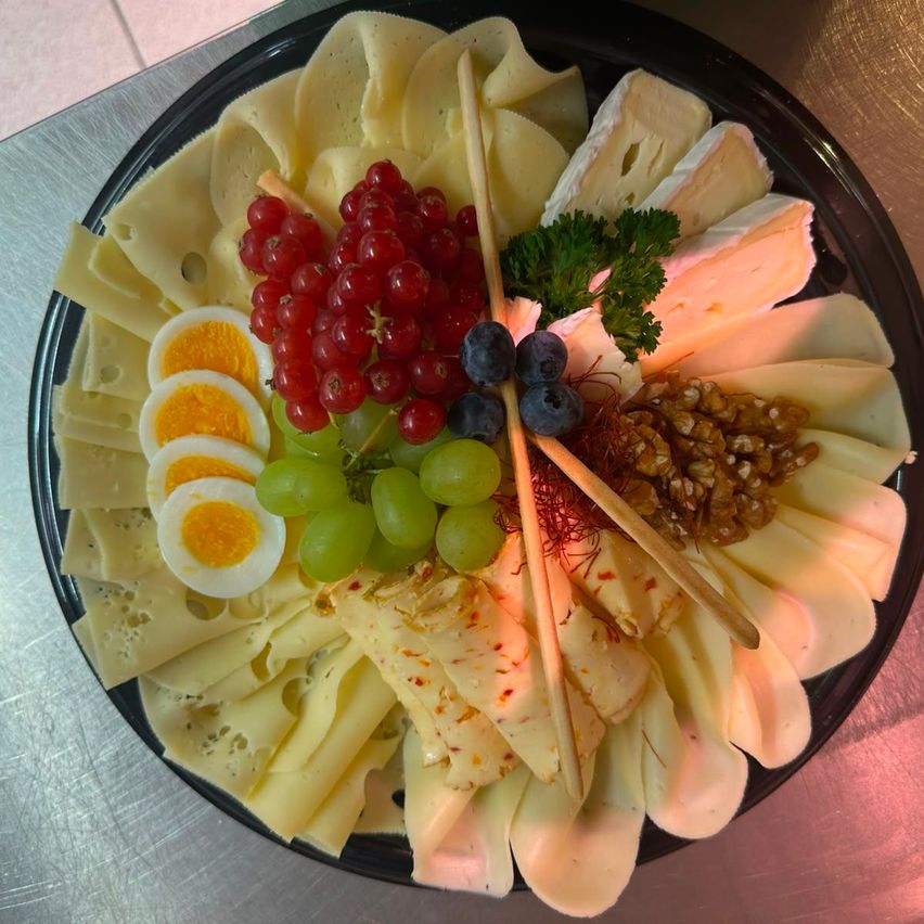 A black plate displays a variety of cheeses, including slices of white cheese, a few hard-boiled eggs, red and green grapes, blueberries, and a sprig of parsley.