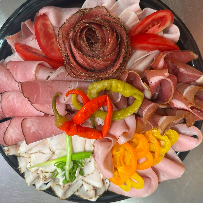 A platter of assorted meats, including sliced ham and salami, arranged with colorful peppers and tomatoes. A rose-shaped piece of meat adds a decorative touch.