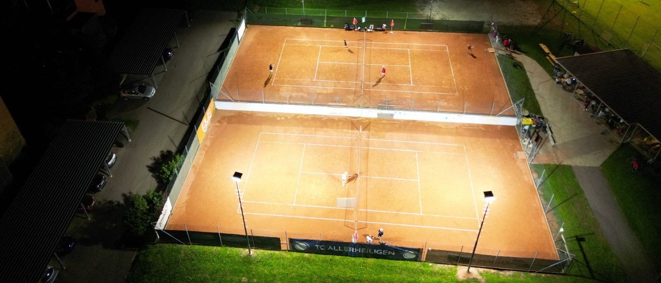 Luftaufnahme eines Tennisplatzes bei Nacht mit Spielern und Zuschauern, beleuchtet von Lampen, in einem Wohngebiet.