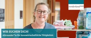 Eine lächelnde Frau mit Brille steht drinnen, vermutlich in einem Laden, mit einem blauen Schild, auf dem 'CHEN DICH!' steht. Sie ist von Gegenständen wie gefalteten Kleidern und einem Regal umgeben. Hinter ihr befindet sich eine rote Wand mit einem weißen Rand.