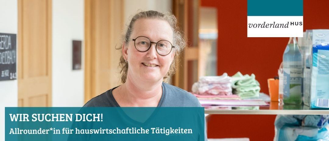 Eine lächelnde Frau mit Brille steht drinnen, vermutlich in einem Laden, mit einem blauen Schild, auf dem 'CHEN DICH!' steht. Sie ist von Gegenständen wie gefalteten Kleidern und einem Regal umgeben. Hinter ihr befindet sich eine rote Wand mit einem weißen Rand.