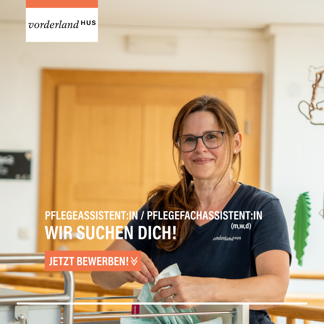Eine Frau mit Brille, die ein Taschentuch hält, lächelt vor einer Tür mit dem Text 'JETZT BEWERBEN!' Dahinter eine Holzwand mit dem Text 'Pflegeassistent:in/Pflegefachassistent:in (m,w,d)'.