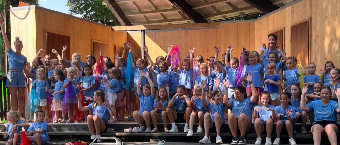 Eine Gruppe von Kindern in blauen T-Shirts steht auf einer Bühne unter einem Holzdach. Sie heben die Hände und lächeln, einige halten rosa und blaue Bänder.