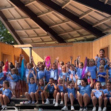 Eine Gruppe von Kindern in blauen T-Shirts, die auf einer Bühne sitzen und stehen, winken und lächeln mit erhobenen Händen. Die Bühne ist aus Holz mit einem Dach, und es gibt Bäume im Hintergrund.