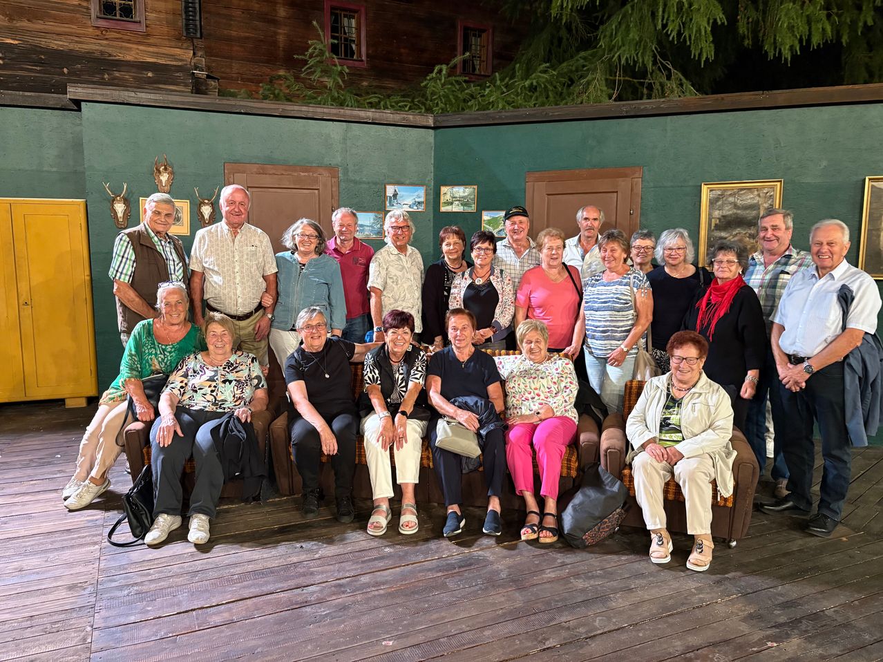 Eine Gruppe älterer Menschen posiert für ein Foto in einem Raum mit Holzböden und grünen Wänden. Einige sitzen auf Sofas, während andere stehen.
