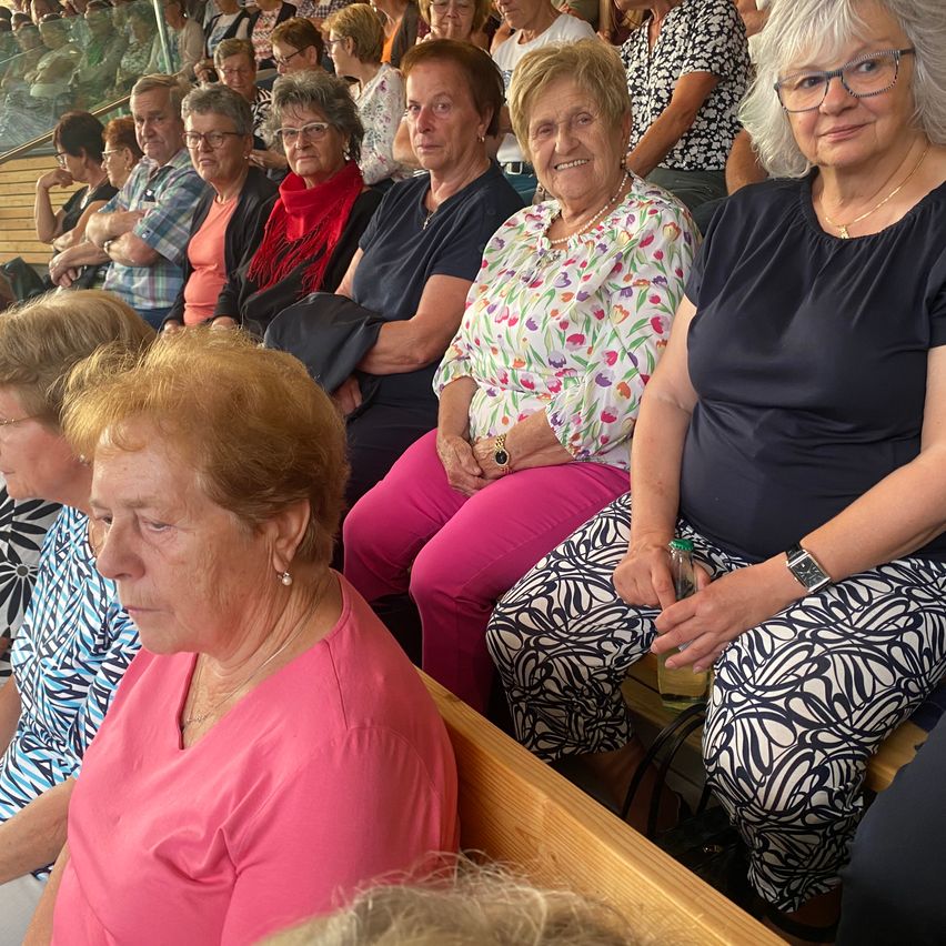 Eine Gruppe älterer Frauen sitzt in einem Stadion, einige tragen Brillen und Halsketten, eine hält ein Getränk.
