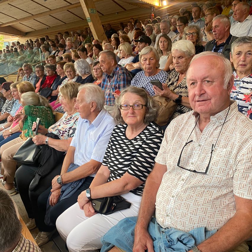 Eine Gruppe älterer Menschen sitzt in einem Stadion, einige mit Brille und halten Handtaschen. Ein Mann mit einer Brille auf seinem Hemd schaut nach vorne.